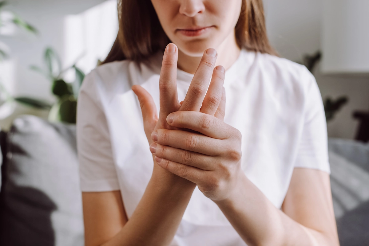 A person in a white shirt sits indoors, holding and massaging their right hand with a concerned expression.