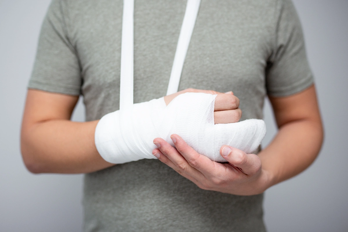A person in a gray shirt holds their bandaged left hand, wrist and arm in a sling. The image conveys a sense of injury and care.