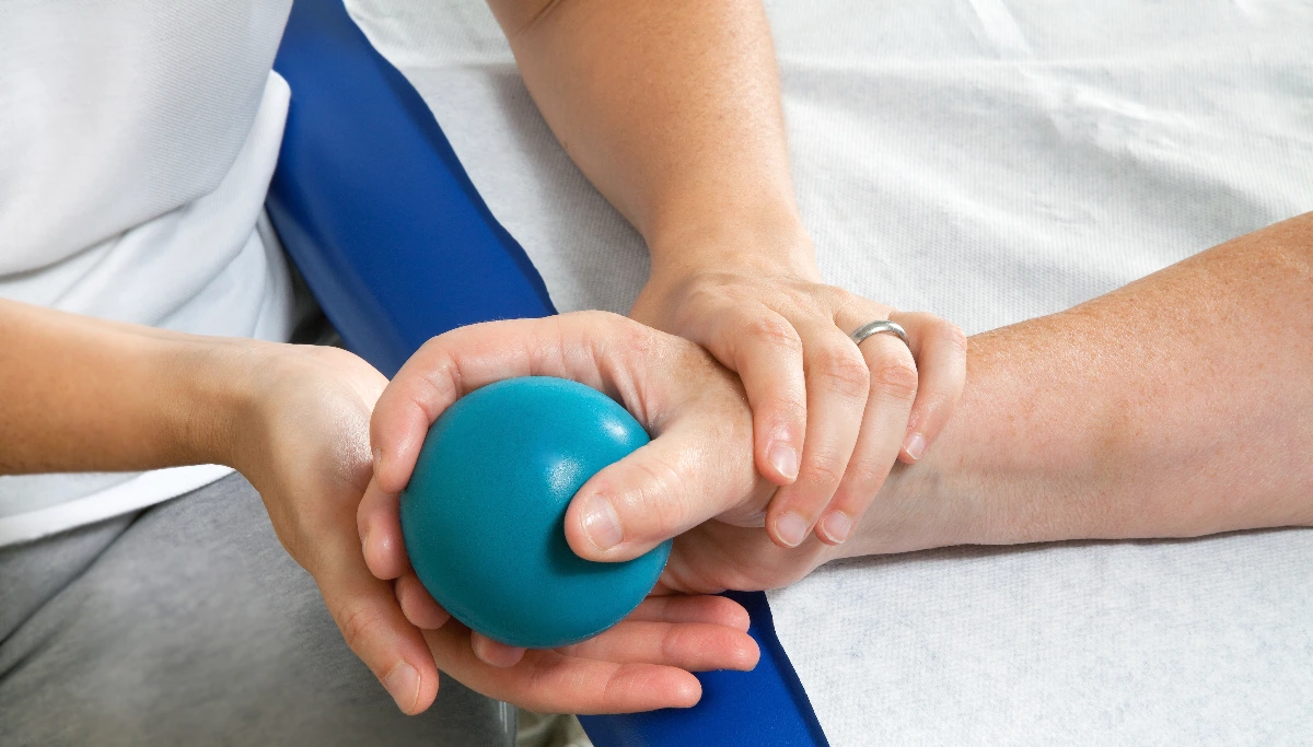 A therapist assists a person with hand exercises using a teal stress ball on a white surface. The scene conveys care and rehabilitation focus on grip strength.