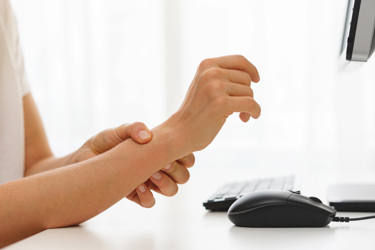 A person seated at a desk gently massages their wrist, possibly indicating discomfort. Nearby are a computer mouse and keyboard, suggesting an office environment.