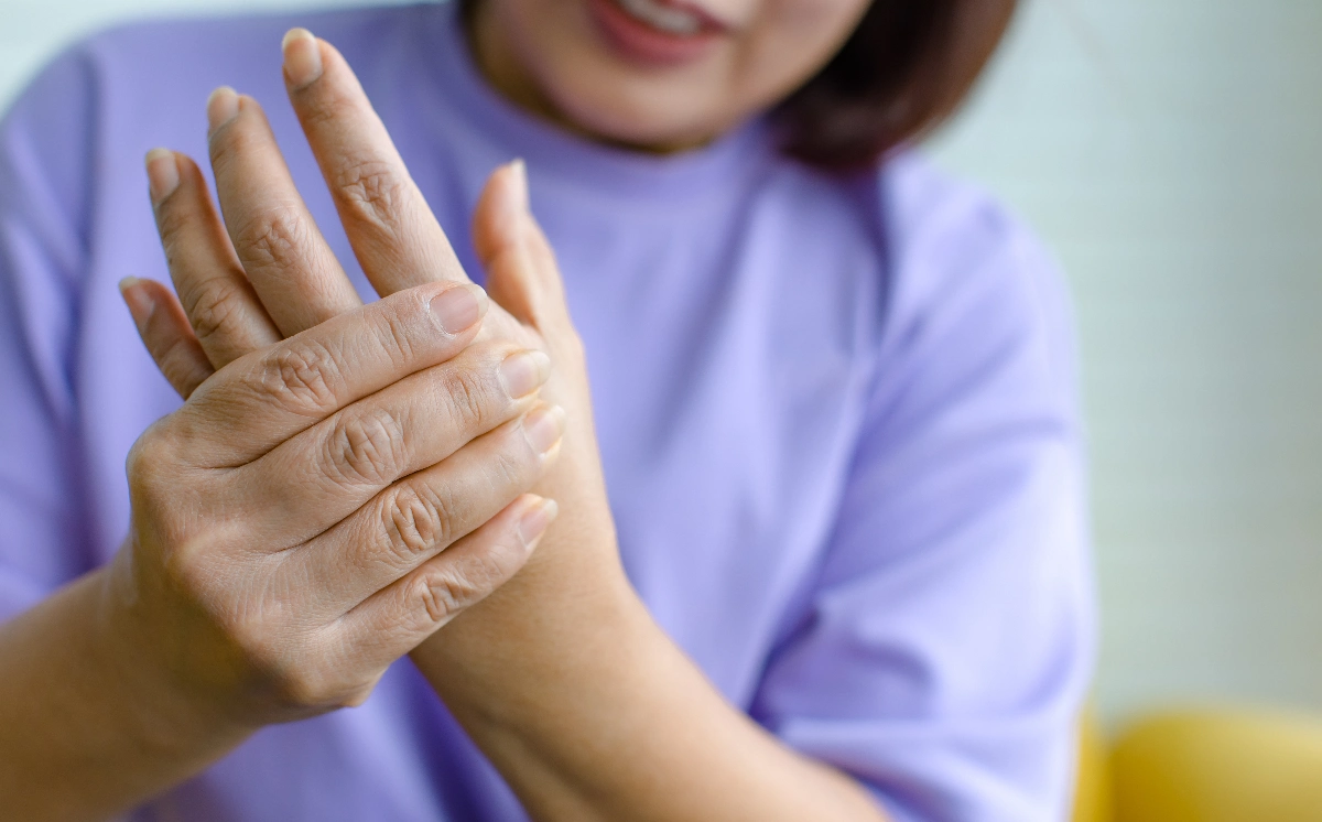 A person wearing a purple shirt holds their right hand with their left, possibly indicating pain or discomfort. The background is softly blurred.