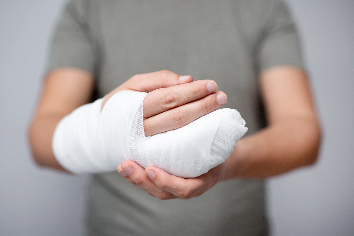 A person in a gray shirt holding their white bandaged right hand with their left hand, conveying care and recovery, set against a plain background.