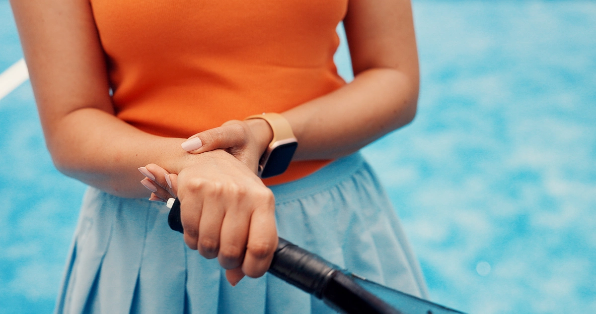 A person grips a tennis racket on a bright blue court. They wear an orange top, light blue skirt, and a smartwatch, conveying a sporty, focused mood. DRUJ Reconstruction Surgeon in Vancouver Dr. Rod French