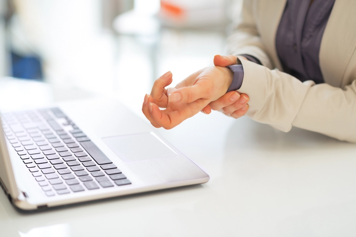 A person in a suit rubs their wrist, suggesting discomfort or strain, next to an open laptop on a bright, clean desk, implying an office setting. Hand & Wrist Ligament Repair Surgeon Dr. Rod French
