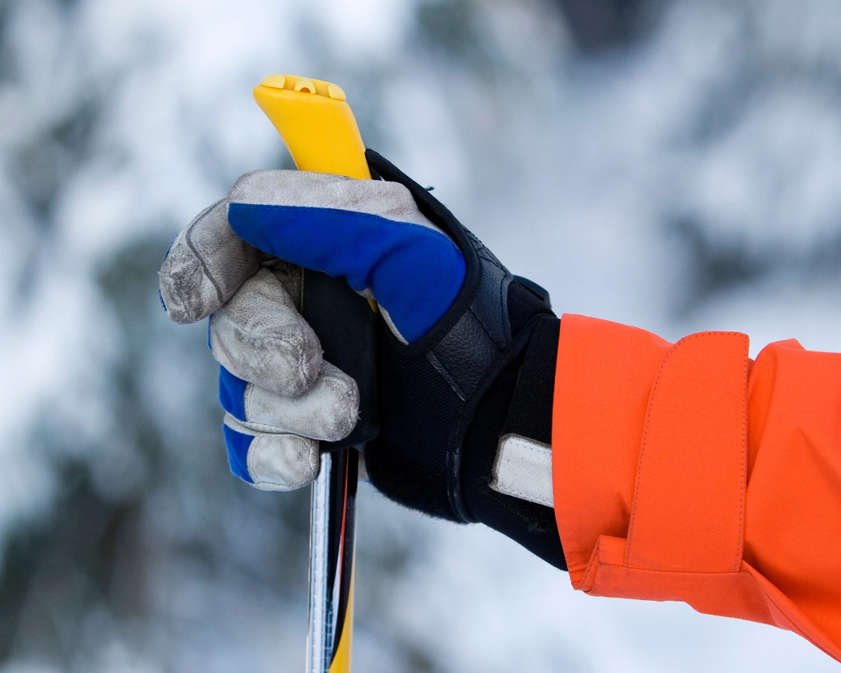 Close-up of a hand in a blue and gray glove gripping a yellow ski pole against a snowy background. The person is wearing an orange jacket, conveying a sense of winter activity. Skier's Thumb (UCL) Repair Surgeon Dr. Rod French Vancouver
