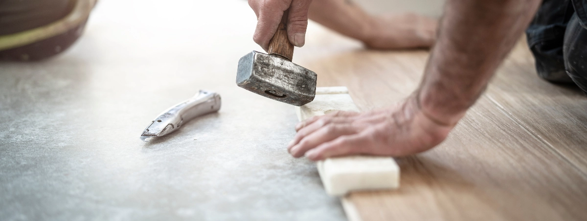Close-up of a person installing flooring, pressing down a board with one hand and using a mallet in the other; a utility knife is on the floor nearby.