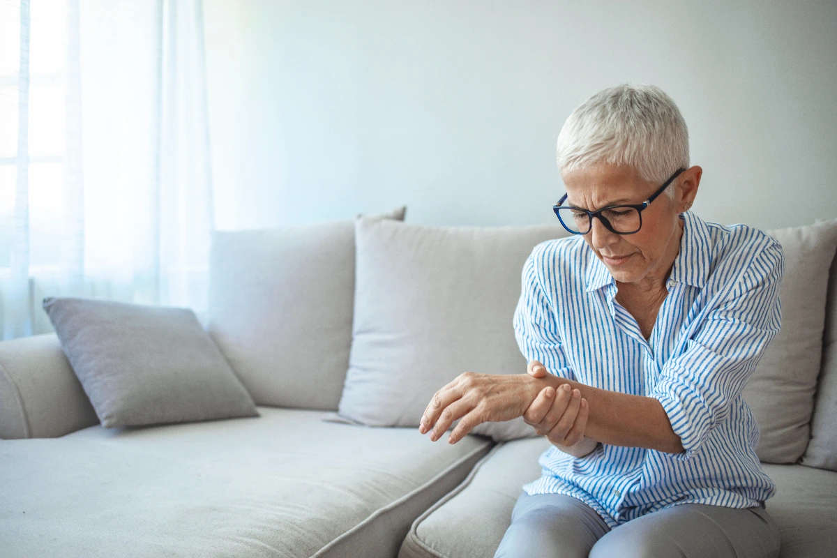 An elderly woman with short white hair sits on a beige sofa, holding her wrist in discomfort. She wears glasses and a striped shirt, looking pained. Wrist Salvage & Fusion Surgery Dr. Rod French Vancouver